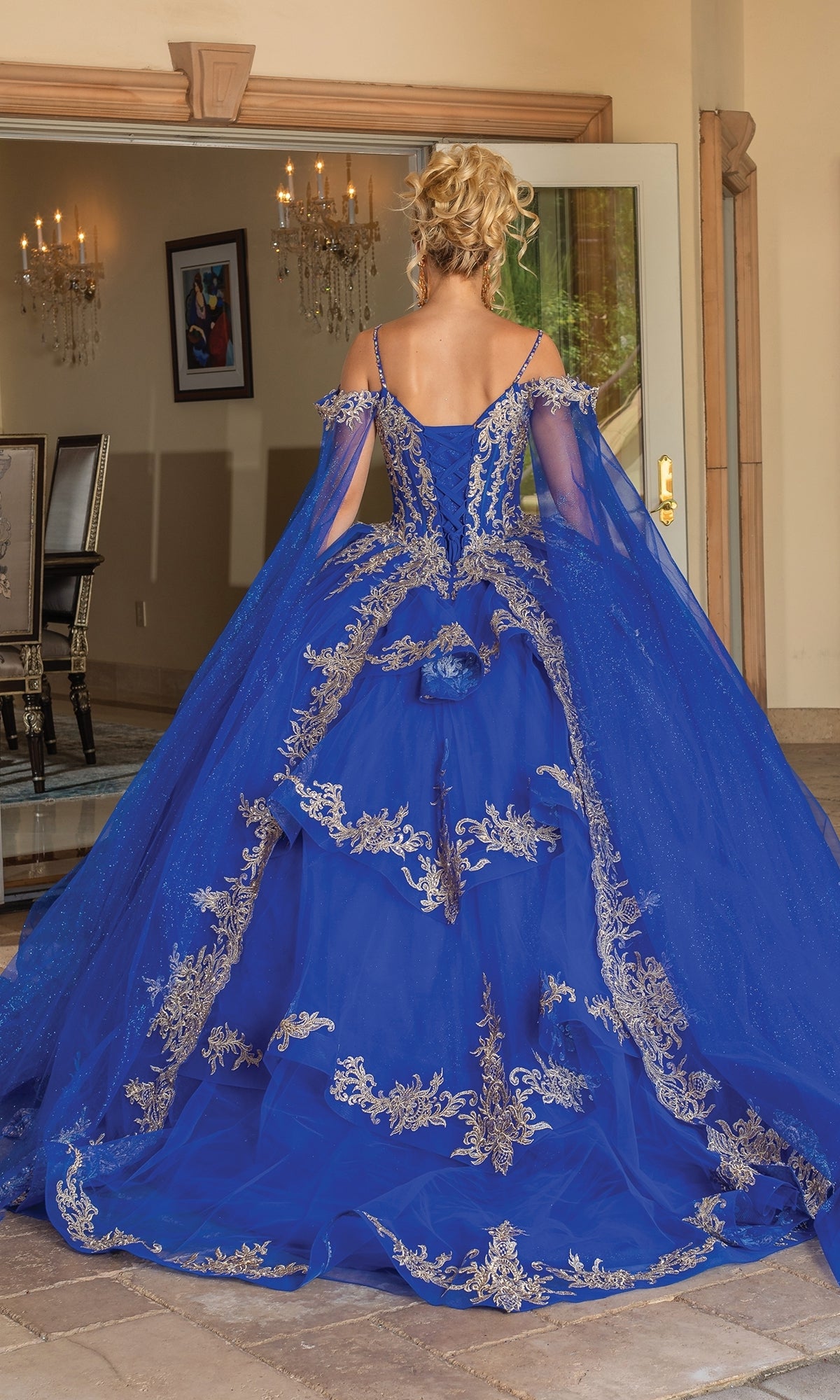 A woman with blond hair in an updo wears the Quinceanera Dress 1768 by Dancing Queen, featuring ornate gold embroidery and cape sleeves, as she stands with her back to the camera in an elegant room.